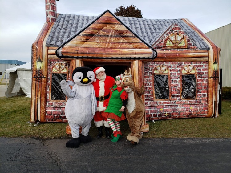 Santa, elves, a penguin, and a reindeer outside an inflatable Santa house at a community event